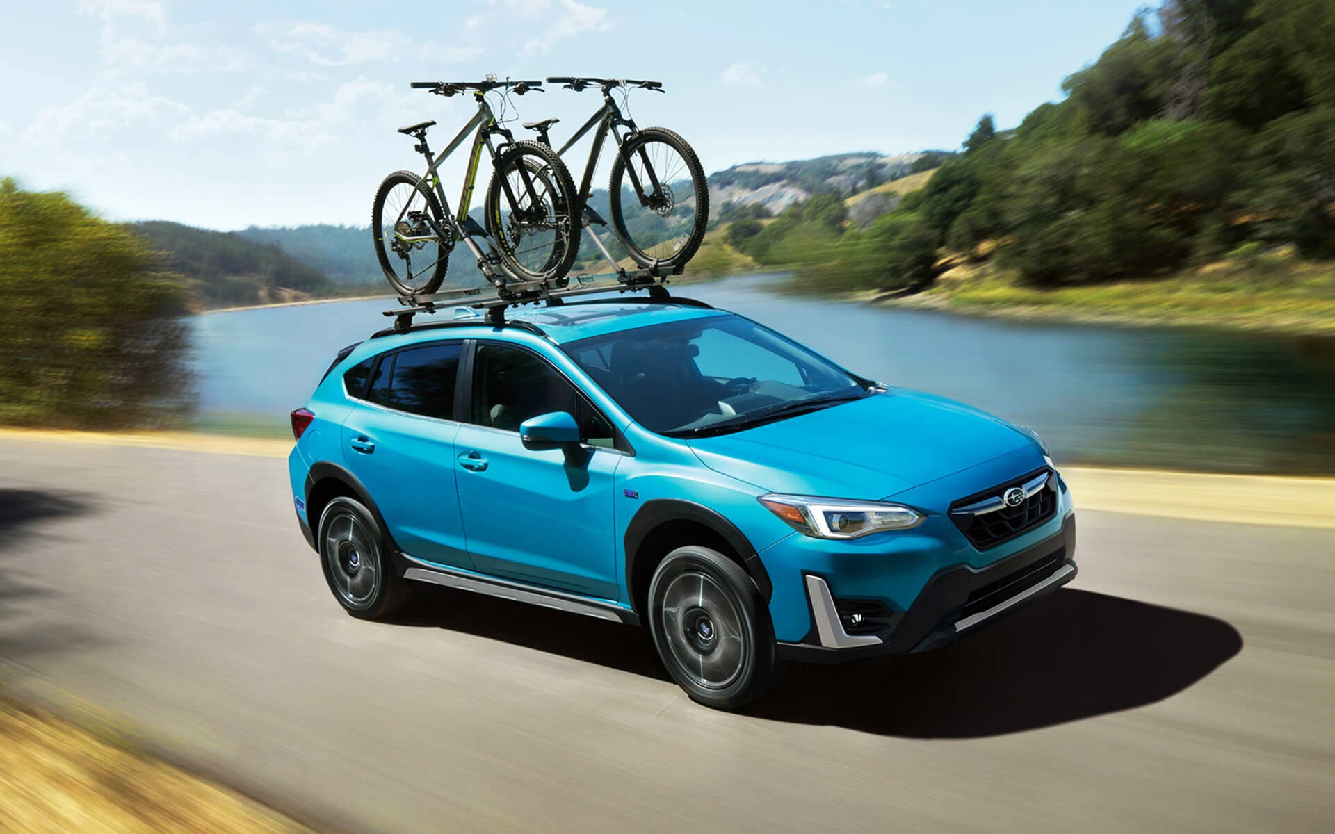 A blue Crosstrek Hybrid with two bicycles on its roof rack driving beside a river | Subaru Superstore of Surprise in Surprise AZ